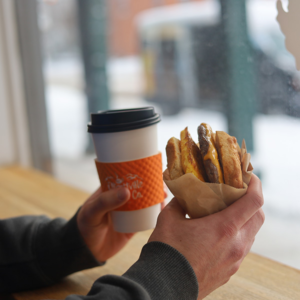 man holding coffee with signature orange coffee sleeve and in the other hand holding a sausage egg breakfast sandwich in the front window