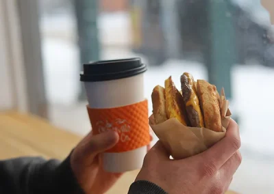 Hand holding a coffee cup with an orange sleeve and a breakfast sandwich
